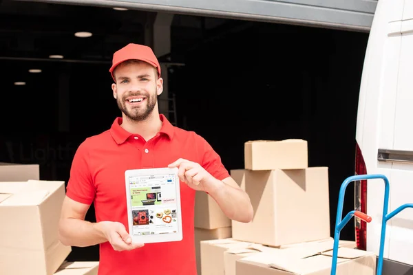 Retrato del repartidor sonriente mostrando la tableta con el sitio web de ebay en la pantalla en las manos con la carga detrás - foto de stock