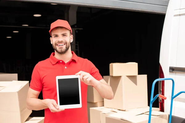 Portrait of smiling delivery man showing tablet with blank screen in hands with cargo behind — Stock Photo