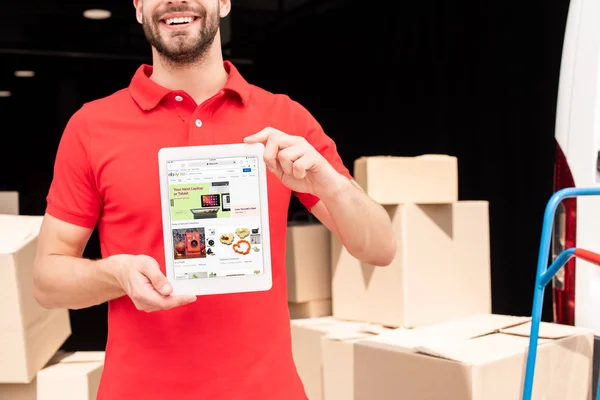 Cropped shot of smiling delivery man showing tablet with ebay website on screen in hands with cargo behind — Stock Photo