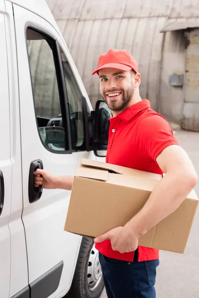 Homem de entrega sorridente em uniforme com caixa de papelão em pé perto de van branca na rua — Fotografia de Stock