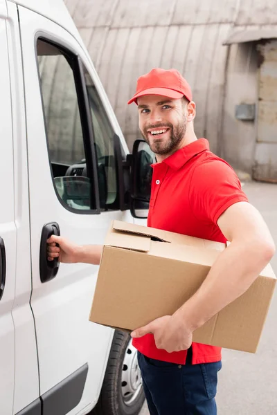 Livreur souriant en uniforme avec boîte en carton debout près de van blanc dans la rue — Photo de stock