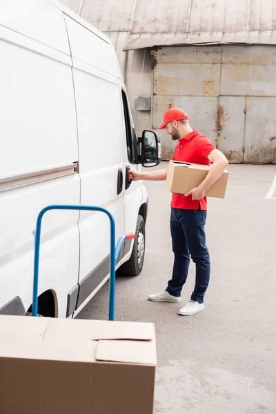 Side view of young delivery man in uniform with cardboard box standing near white van in street — Stock Photo