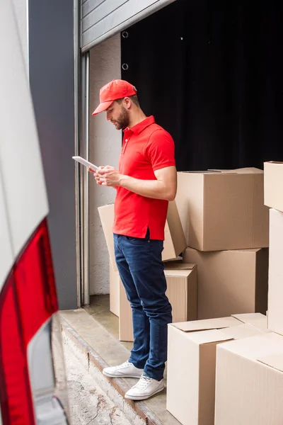 Side view of delivery man in red uniform using digital tablet with cargo behind — Stock Photo