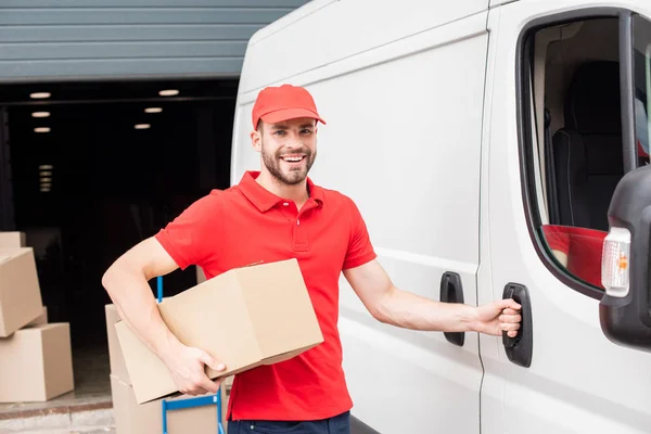 Portrait of smiling delivery man in uniform with cardboard box standing near white van in street — Stock Photo