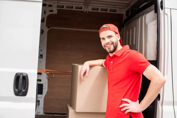 Smiling young delivery man in uniform standing near van with cargo — Stock Photo