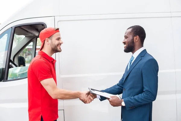 Homem de entrega sorridente e empresário afro-americano apertando as mãos perto de van — Fotografia de Stock