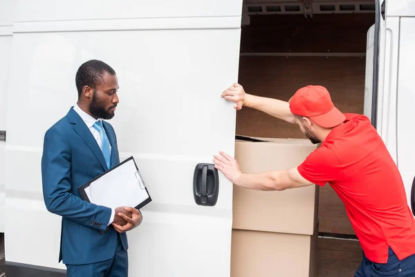 Afro-américain homme d'affaires avec bloc-notes regardant caucasien livreur fermeture van avec cargo — Photo de stock