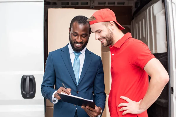 Portrait of smiling multicultural businessman and delivery man looking at notepad — Stock Photo