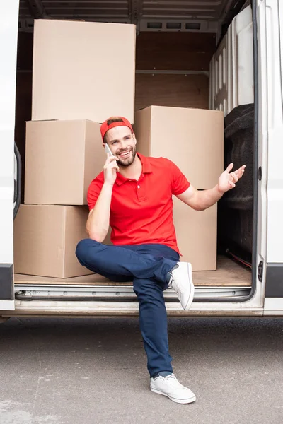 Улыбающийся курьер в красной форме разговаривает по смартфону, отдыхая в фургоне с картонными коробками — стоковое фото