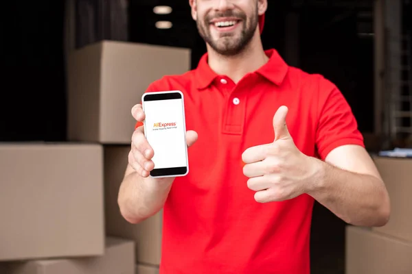 Vista parcial del hombre alegre de la entrega con el teléfono inteligente con el logotipo de aliexpress en la pantalla que muestra el pulgar hacia arriba - foto de stock
