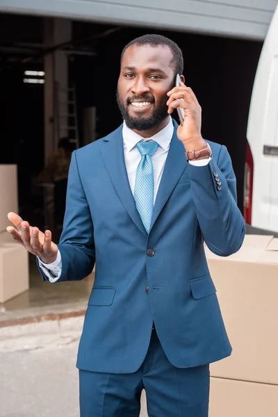 Retrato de sorridente empresário afro-americano falando em smartphone com caixas de papelão atrás — Fotografia de Stock