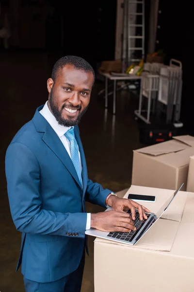 Vue latérale de souriant homme d'affaires afro-américain avec ordinateur portable sur boîte en carton — Photo de stock