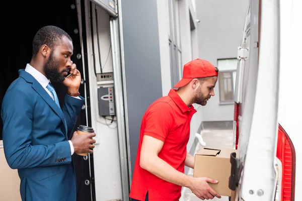 African american businessman with coffee to go controlling caucasian delivery man while talking on smartphone — Stock Photo