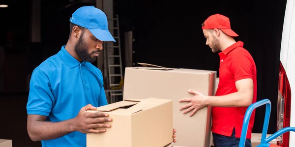 Vista lateral de los repartidores multiétnicos en uniforme rojo y azul con cajas de cartón - foto de stock