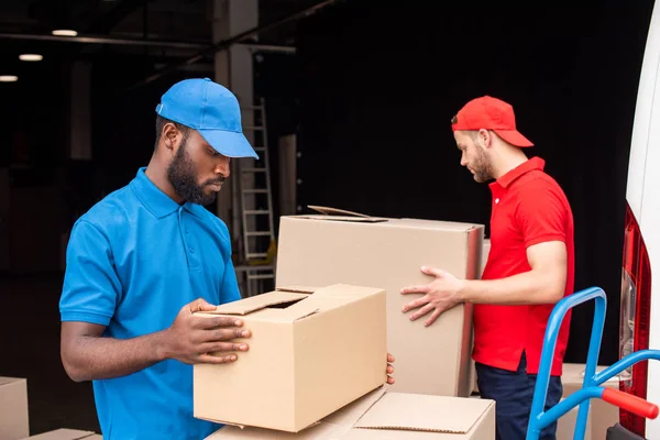 Vista lateral de homens de entrega multicultural em uniforme vermelho e azul com caixas de papelão — Fotografia de Stock