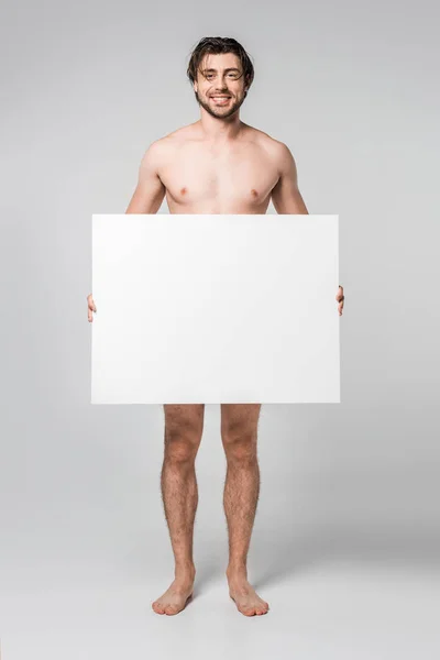 Sonriente guapo hombre desnudo sosteniendo banner en blanco sobre fondo gris - foto de stock