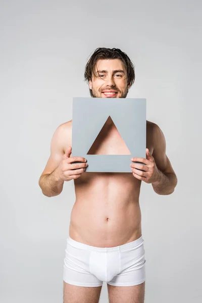 Portrait de jeune homme souriant en sous-vêtements tenant triangle figurine en papier isolé sur gris — Photo de stock