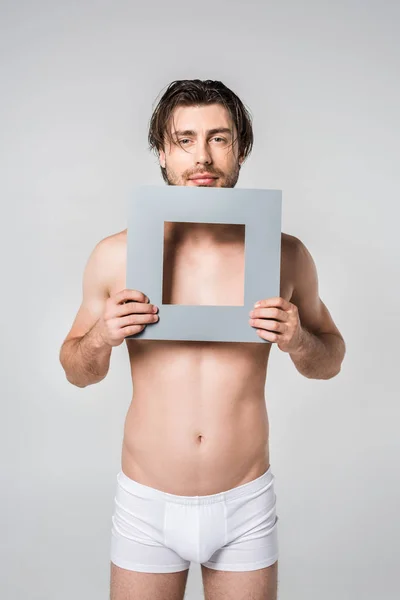 Портрет молодого человека в нижнем белье, держащего квадратную бумажную фигуру, изолированную на сером — стоковое фото