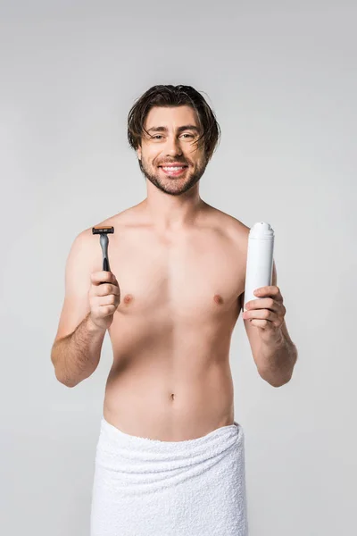 Портрет улыбающегося мужчины в белом полотенце с бритвой и пеной для бритья в руках, изолированных на сером — стоковое фото