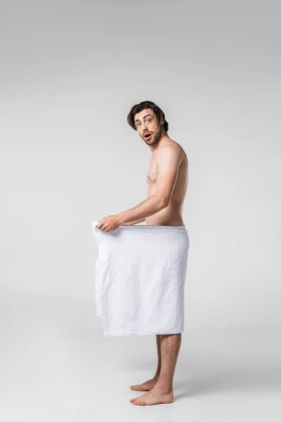 Vista lateral do homem chocado em toalha branca olhando para a câmera no fundo cinza — Fotografia de Stock