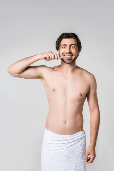 Ritratto di uomo allegro in asciugamano bianco con spazzolino isolato su grigio — Foto stock