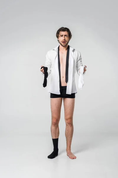 Shocked young man in shirt and underwear holding black and white socks on grey backdrop — Stock Photo