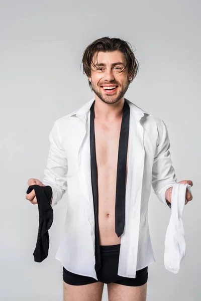 Portrait of smiling young man in shirt and underwear holding black and white socks isolated on grey — Stock Photo