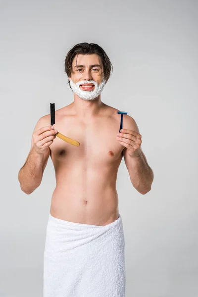 Ritratto di uomo sorridente in asciugamano bianco con schiuma da barba sul viso e rasoi in mani isolate su grigio — Foto stock