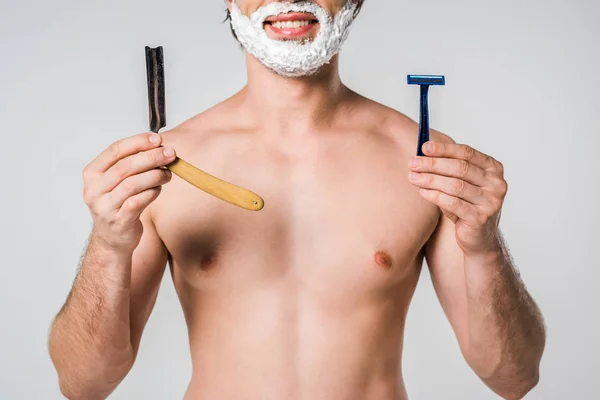 Vista parcial del hombre sin camisa con espuma de afeitar en la cara que sostiene las máquinas de afeitar aisladas en gris - foto de stock