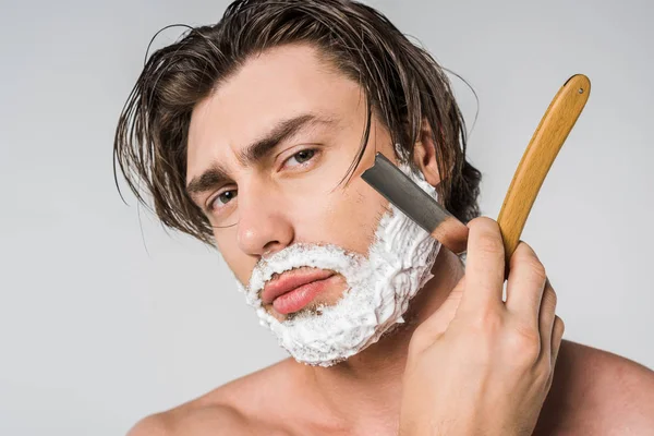 Portrait of shirtless man with foam on face shaving beard with straight razor isolated on grey — Stock Photo