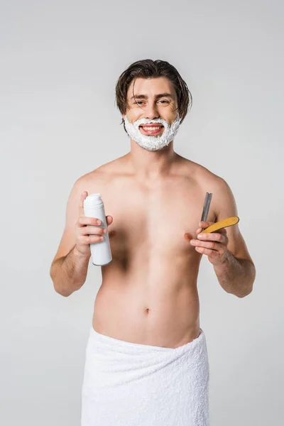 Ritratto di uomo sorridente in asciugamano bianco con schiuma da barba sul viso e rasoio dritto in mano isolato su grigio — Foto stock