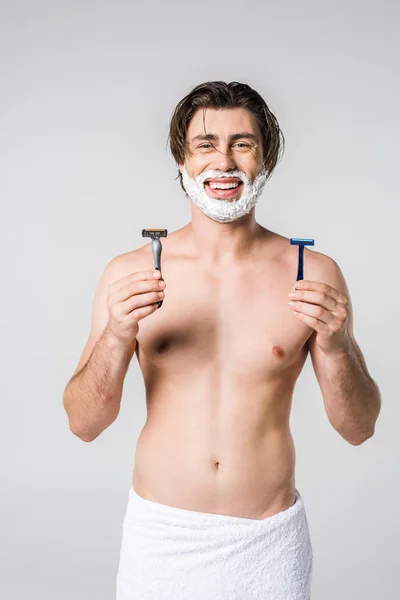 Portrait of happy man in white towel with shaving foam on face and razors in hands isolated on grey — Stock Photo