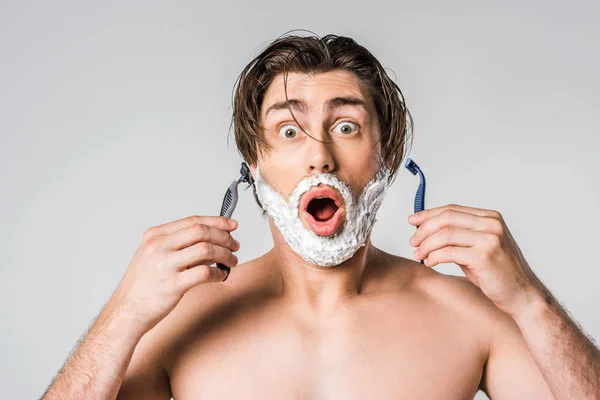 Retrato del hombre sorprendido con espuma de afeitar en la cara que sostiene las máquinas de afeitar aisladas en gris - foto de stock