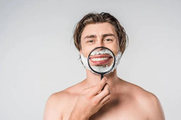 Portrait of smiling man with shaving foam on face and magnifying glass isolated on grey — Stock Photo