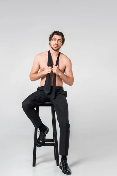 Мужчина без рубашки в черных штанах с галстуком сидит на стуле на сером фоне — стоковое фото
