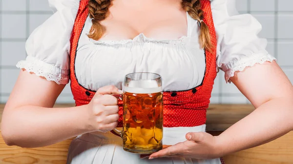 Imagen recortada de la camarera oktoberfest en vestido alemán tradicional sosteniendo taza de cerveza ligera cerca del mostrador del bar - foto de stock
