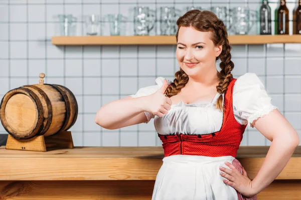 Oktoberfest waitress in traditional bavarian dress doing thumb up gesture and standing near bar counter with beer barrel — Stock Photo