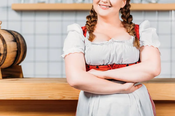 Imagen recortada de la camarera oktoberfest en vestido bavariano tradicional con brazos cruzados de pie cerca del mostrador del bar con barril de cerveza - foto de stock