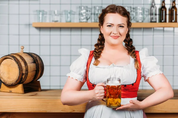 Smiling oktoberfest bartender in traditional bavarian dress showing mug of light beer near bar counter — Stock Photo