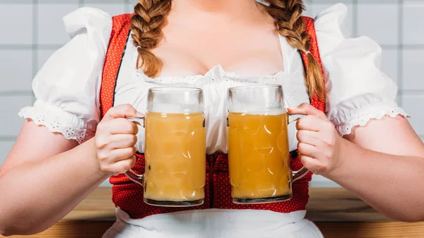 Imagem cortada de garçonete oktoberfest em vestido tradicional bávaro segurando canecas de cerveja leve com espuma perto do balcão bar — Fotografia de Stock