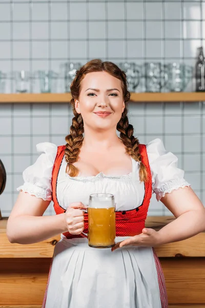 Smiling oktoberfest waitress in traditional bavarian dress showing mug of light beer near bar counter — Stock Photo