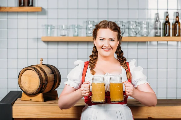 Camarera sonriente oktoberfest en vestido alemán tradicional sosteniendo tazas de cerveza ligera cerca del mostrador del bar - foto de stock