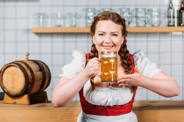 Heureuse serveuse oktoberfest en robe bavaroise traditionnelle sentant la bière légère près du comptoir du bar — Photo de stock