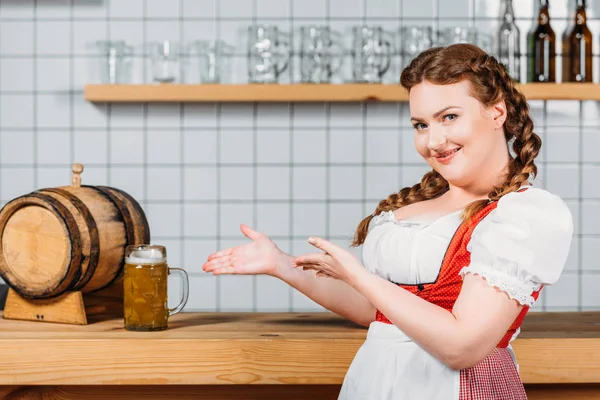 Heureuse serveuse oktoberfest en robe bavaroise traditionnelle pointant du doigt une tasse de bière légère sur le comptoir du bar — Photo de stock