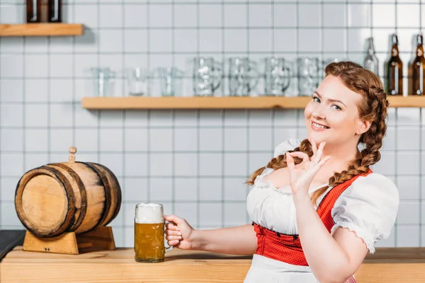 Oktoberfest waitress in traditional bavarian dress doing ok gesture and standing near bar counter with light beer — Stock Photo