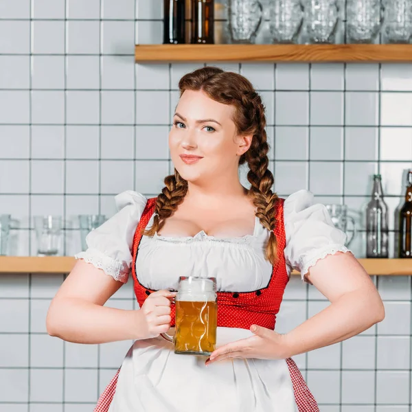 Beautiful oktoberfest waitress in traditional bavarian dress showing at mug of light beer near bar counter — Stock Photo