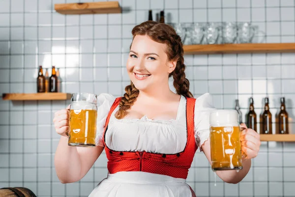 Happy oktoberfest waitress in traditional bavarian dress showing mugs of light beer near bar counter — Stock Photo