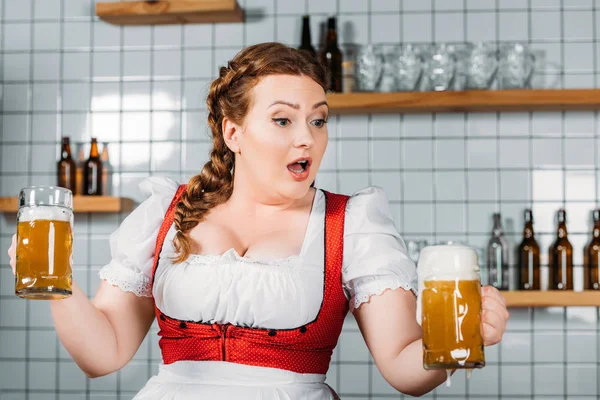 Choquée serveuse oktoberfest en robe bavaroise traditionnelle tenant des tasses avec de la bière moussée qui coule dans le bar — Photo de stock