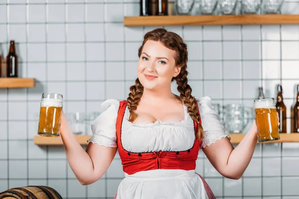 Sorrindo oktoberfest garçonete em vestido tradicional bávaro segurando canecas de cerveja leve no bar — Fotografia de Stock