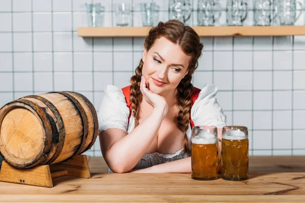 Sorrindo oktoberfest garçonete em vestido tradicional alemão de pé no balcão de bar com barril de cerveja e duas canecas de cerveja leve — Fotografia de Stock